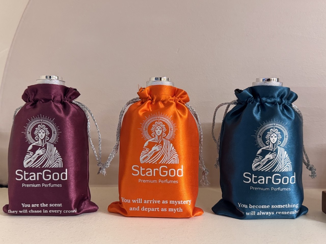 Three drawstring satin pouches in aubergine, orange, and teal, each printed with a line in cream.